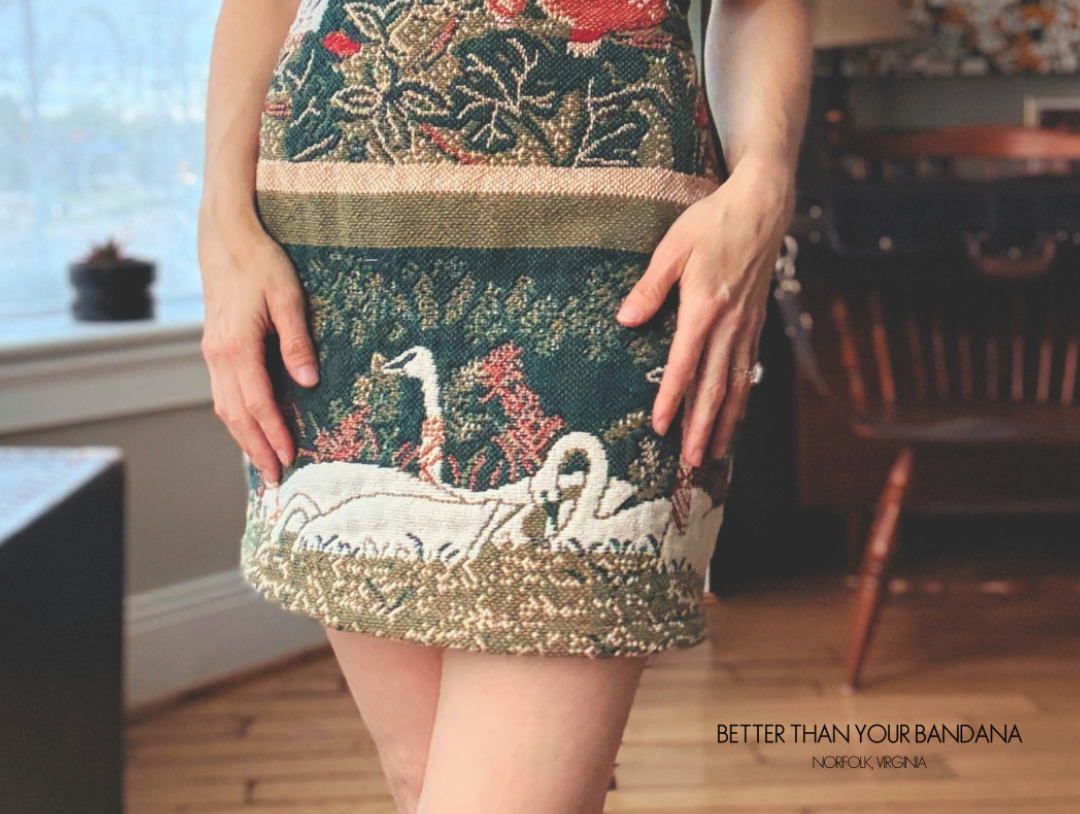 A woman wearing a sleeveless mini dress with an intricate design featuring birds, flowers, and swans on a dark green background.